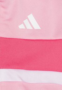 Adidas audums rozā toņos ar teksturētām rakstām; augšējā kreisajā stūrī ir balts logotips, kas gludi kontrastē ar ribotu fona.