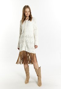 Vit, strukturerad cardigan med geometriska mönster, som har en fransig, tanfärgad nederkant. Matchad med tanfärgade, spetsiga cowboyboots.