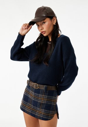 Granatowy sweter dziany z dekoltem w kształcie litery V, w zestawieniu z mini spódnicą w kratę z żółtymi i białymi liniami, z secured brązowym paskiem. Założona brązowa czapka.