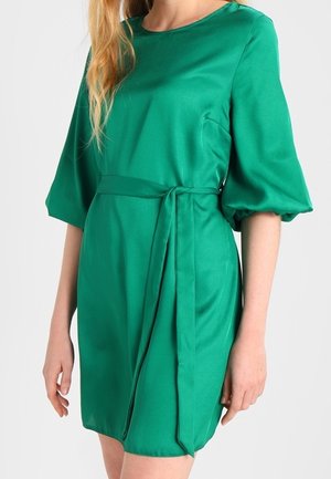 Vrouw draagt een knielange smaragdgroene jurk met pofdriekwartmouwen en een bijpassende stoffen riem gestrikt in de taille.
