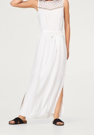 Biała sukienka maxi bez rękawów z koronkową górą, paskiem w talii i bocznym rozcięciem, noszona z czarnymi sandałami wsuwanymi.