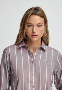 Camicia a righe con bottoni in mauve chiaro e bianco, caratterizzata da un colletto a punta, maniche lunghe e un piccolo logo ricamato sul petto.