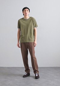 T-shirt verde oliva con bordi bianchi, abbinata a pantaloni marroni e sneakers scure. Design semplice, maniche corte e vestibilità rilassata.