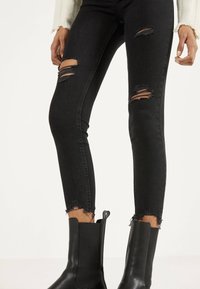 Jeans ajustados negros con rodillas rasgadas, dobladillos deshilachados y una textura suave. Combinados con botas negras hasta el tobillo de diseño elegante.