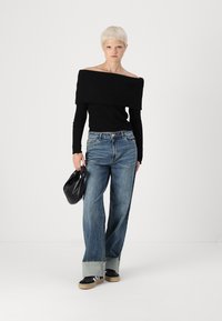 Schwarzes, geripptes Off-Shoulder-Oberteil, hochtaillierte hellblaue Jeans mit umgekrempelten Säumen, schwarze Schuhe, hält eine schwarze Tasche, einfarbiger Hintergrund.
