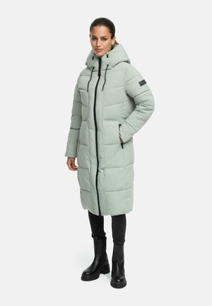 Manteau matelassé vert clair avec un col haut et une fermeture éclair, featuring un zip noir, des poches latérales et une capuche. Porté avec des bottes noires et un pantalon.