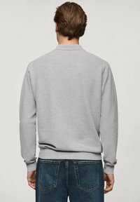 Mann mit kurzen braunen Haaren, der einen hellgrauen, langärmeligen Pullover und blaue Jeans trägt und mit dem Rücken zu einer schlichten Hintergrund steht.