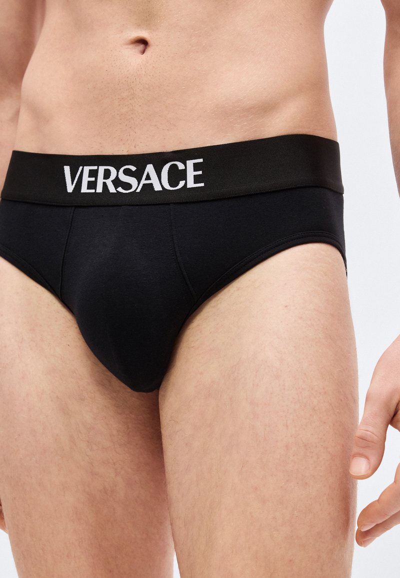 Man wearing black Versace briefs with logo on waistband, shown from waist to upper thighs against plain background.