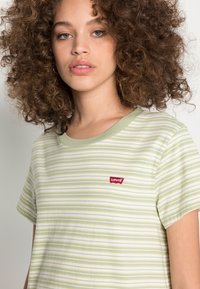 Jovem mulher com cabelo encaracolado, vestindo uma t-shirt da Levi's em verde claro e branca com riscas, olhando ligeiramente para além da câmara num fundo simples.