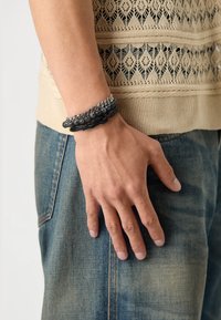 Sorte læder- og sølvkædearmbånd med perle-detaljer, båret på en hånd mod en tekstureret lys beige strikket top og denim jeans.