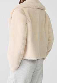 Jacke aus cremefarbenem Kunstpelz mit weitem Kragen, lockerem Schnitt und weicher Textur. Mit glatten Nähten und kurzem Schnitt. Graue Hose sichtbar.