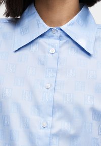Világoskék ing finom, egész felületén elhelyezett logómintával, klasszikus gallérral és gombos záródással. Simogató, puha textúrájú anyag.