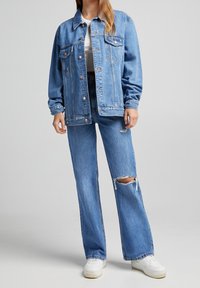Light blue denim jacket with a relaxed fit, front pockets, and copper buttons, paired with matching wide-leg ripped jeans. White sneakers.