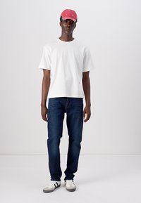 T-shirt bianco in cotone, jeans blu scuro e un cappellino da baseball rosso con dettagli neri. Le calzature hanno una base bianca con strisce nere e suole grigie.