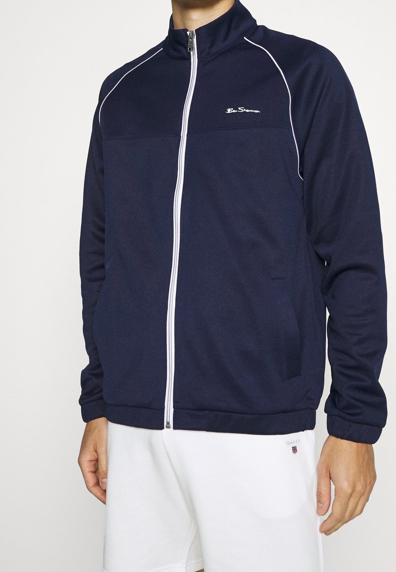 Navy zip-up track jacket with white piping, high collar, and ribbed cuffs; features logo on the chest and side pockets.