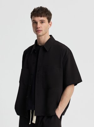 Młody mężczyzna z krótkimi brązowymi włosami, noszący luźną czarną koszulę z dużymi przednimi kieszeniami, stoi na jednolitym jasnym tle.