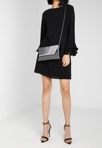 Robe noire avec des manches évasées, assortie à une pochette grise avec un accent métallique, et des escarpins noirs à lanières. Tissu lisse.