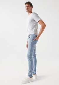 Jeans de mezclilla azul claro con corte slim, combinados con una camiseta blanca ajustada y zapatillas blancas, de pie contra un fondo liso.