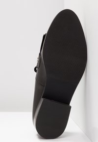 Chaussure habillée en cuir noir, inclinée vers le haut, montrant une semelle texturée et un petit talon bloc sur fond blanc.