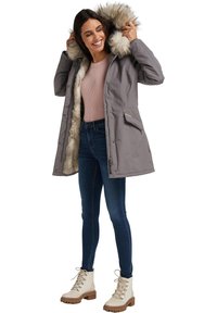 Grijze parka met een capuchon van nepbont, rits- en drukknoopsluitingen, twee zijzakken, gedragen over een lichtroze trui en blauwe jeans, gecombineerd met beige laarzen.