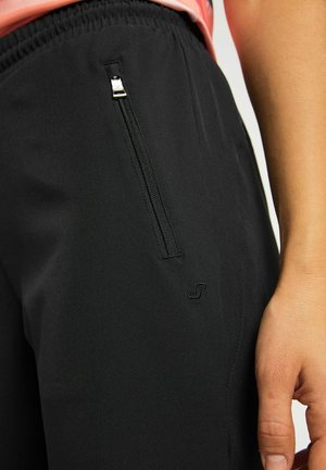 Schwarze Sporthose mit elastischem Bund, ausgestattet mit einer Reißverschlusstasche an der Seite und einem kleinen Logo nahe dem Saum. Weiche, glatte Textur.