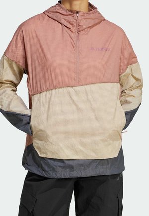 Personne portant un coupe-vent à blocs de couleurs rose, beige et gris, manches longues, fermeture éclair frontale et pantalon noir avec poches.