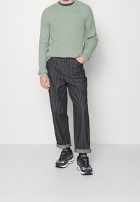 Jasnozielony sweter o teksturze, ciemne spodnie w paski z podwiniętymi nogawkami oraz czarne sneakersy z białymi akcentami.