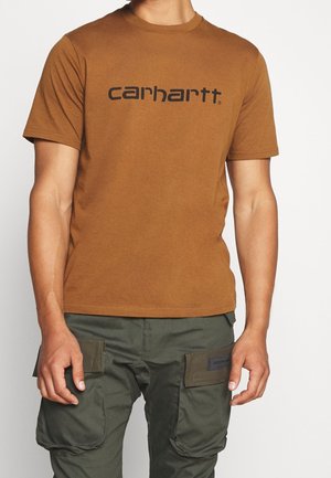Mężczyzna w brązowej koszuli z krótkim rękawem z logo "Carhartt" oraz zielonych spodniach typu cargo z zapinanymi na guziki kieszeniami z przodu.