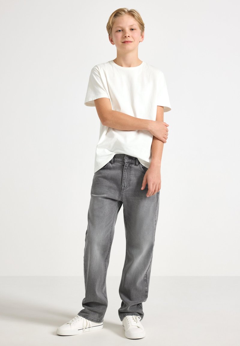 Vit kortärmad T-shirt i kombination med grå denimjeans och vita sneakers, stående mot en ljus bakgrund. Avslappnad passform.