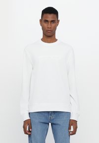 White sweatshirt with ribbed crew neckline and cuffs, featuring an embossed "MICHAEL KORS" logo on the front, paired with light blue jeans.
