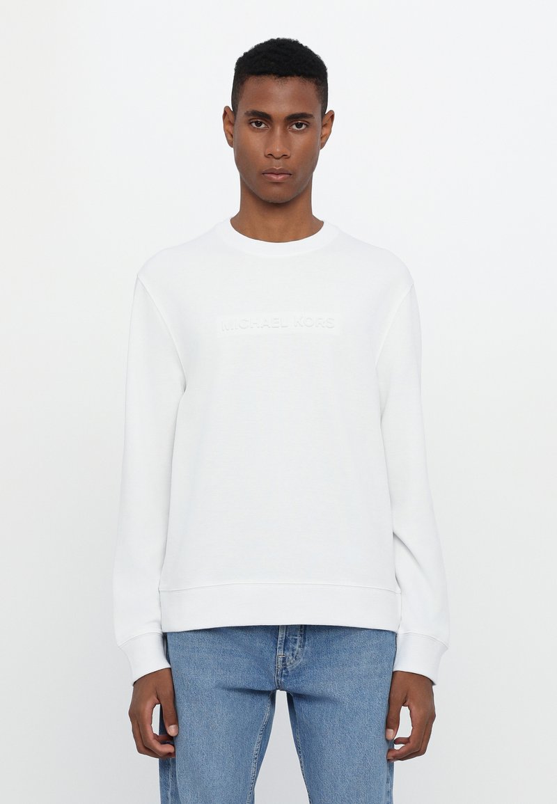 White sweatshirt with ribbed crew neckline and cuffs, featuring an embossed "MICHAEL KORS" logo on the front, paired with light blue jeans.