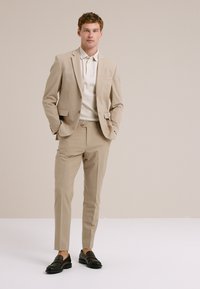 Un joven se presenta con confianza en un traje beige claro, una camiseta polo blanca y zapatos de charol oscuros frente a un fondo neutro.