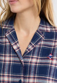 Granatowa koszula w kratę z czerwonymi i białymi wzorami, zapinana na guziki, z haftem przedstawiającym narciarza na obszarze kieszeni na piersi. Wykonana z bawełny.