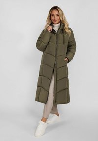 Nainen, joka on pukeutunut pitkään oliivinvihreään untuvatakkiin, beigeihin housuihin ja valkoisiin tennareihin, seisoo vaaleanharmaata taustaa vasten.