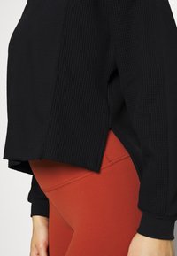 Schwarzes geripptes Langarmoberteil mit seitlichen Schlitzen, kombiniert mit enganliegenden dunkelroten Leggings. Das Oberteil hat einen strukturierten Stoff.