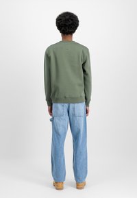 Πράσινη μπλούζα με ριχτές μανσέτες και στρίφωμα, συνδυασμένη με ανοιχτόχρωμο μπλε χαλαρούς τζιν, φανερωμένη από πίσω σε έναν απλό φόντο.