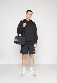 Black hoodie with kangaroo pocket, paired with black shorts. White Adidas logo on shorts and bag. Model holding a black duffel bag.