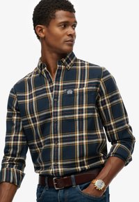 Camisa de cuadros en azul marino con acentos en amarillo, blanco y rojo. Mangas largas, cuello tipo polo, bolsillo en el pecho y etiqueta de marca. Llevada con jeans azules.