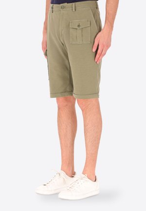 Uomo che indossa pantaloncini cargo verde oliva al ginocchio con tasche con bottoni e scarpe da ginnastica bianche, in piedi davanti a uno sfondo semplice.