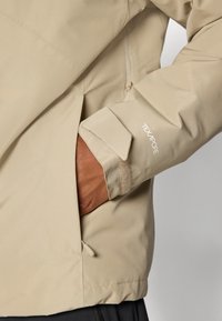 Beige vandtæt jakke med justerbar manchet, lynlåslomme på siden og tekstureret stof. Har logo-detalje på ærmet.
