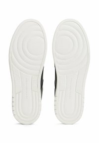 Soli delle sneaker nere con fondi in gomma bianca testurizzata, caratterizzati da motivi circolari e branding. I bordi superiori lisci curvano dolcemente.