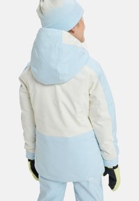 Lichtblauwe en crème skijas met een capuchon. Heeft een kleurblokontwerp, een gladde textuur en zwarte handschoenen met groene accenten.