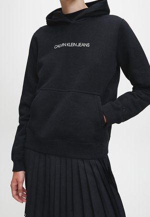Personne portant un sweat à capuche noir Calvin Klein avec poche avant et logo blanc, associé à une jupe plissée noire.