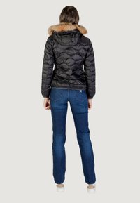 Zwarte pufferjack met een doorgestikt ontwerp en een met bont gevoerde capuchon, gecombineerd met blauwe denim jeans en witte sneakers, gezien vanuit de achterkant.