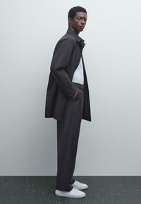 Donker, maatwerk trenchcoat over een wit shirt, gecombineerd met losse grijze broek en witte sneakers. Gladde stof met een gestructureerd ontwerp.