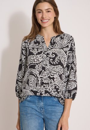 Žena se usmívá, nosí černobílou halenku s paisley vzorem a tříčtvrtečními rukávy a modré džínové kalhoty, stojí před jednoduchým pozadím.