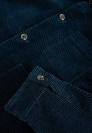 Tmavě modrý kordový materiál s vertikálními pruhy, obsahuje stříbrné knoflíky a náprsní kapsu. Textura je měkká a strukturovaná.