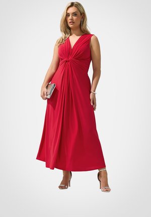 Mujer con un vestido rojo sin mangas hasta el suelo con un nudo en la parte delantera, sosteniendo un bolso de mano plateado y llevando tacones y pendientes plateados.