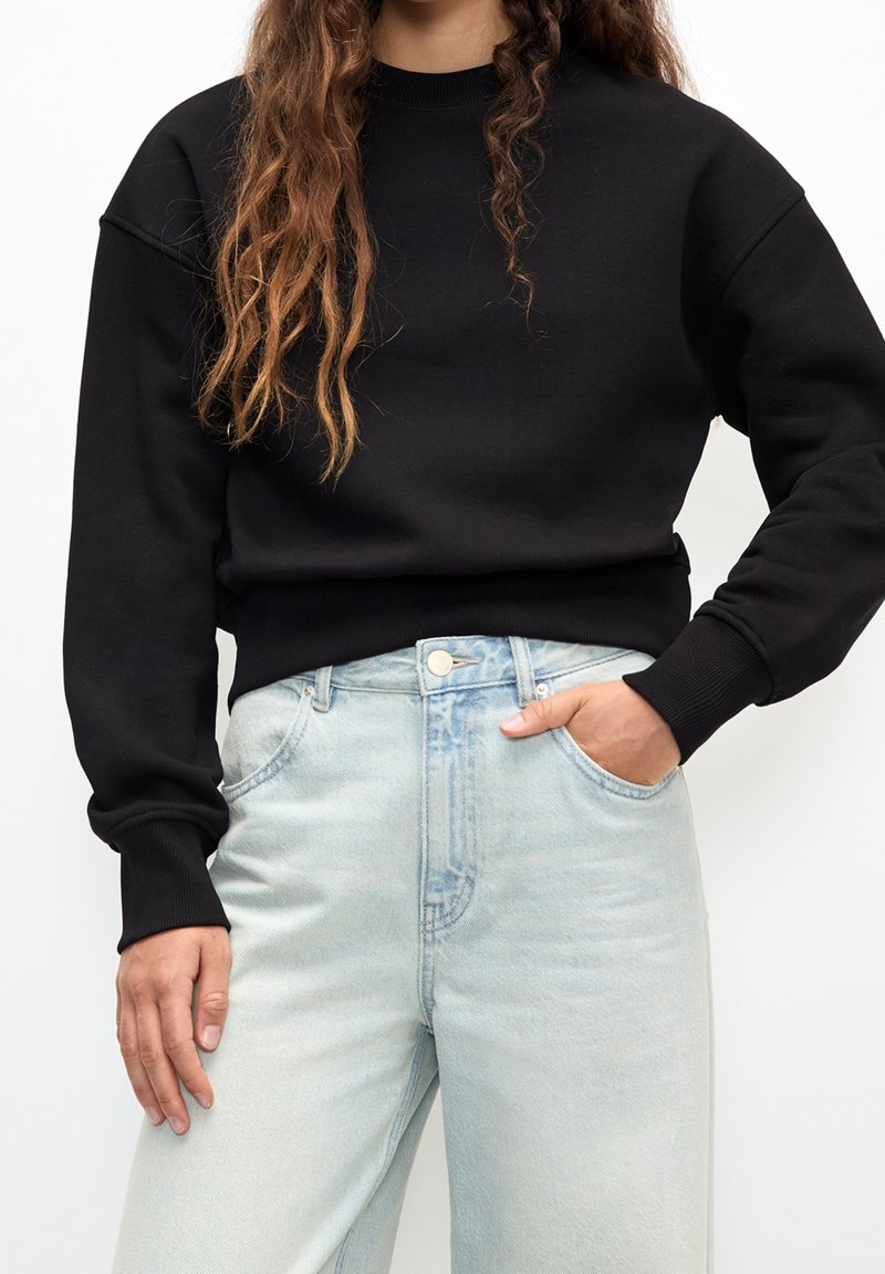 Femme en sweat-shirt noir à manches longues et jean bleu clair taille haute, une main dans la poche, sur fond blanc.