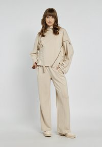 Beżowy oversize'owy sweter z unikalnym diagonalnym wzorem, z zamkami błyskawicznymi i stójką, w połączeniu z szerokimi spodniami i pasującymi butami.